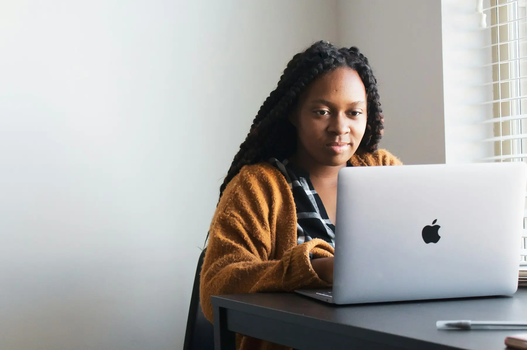 A woman working at an uncluttered desk with a laptop, sitting upright beside a bright window.