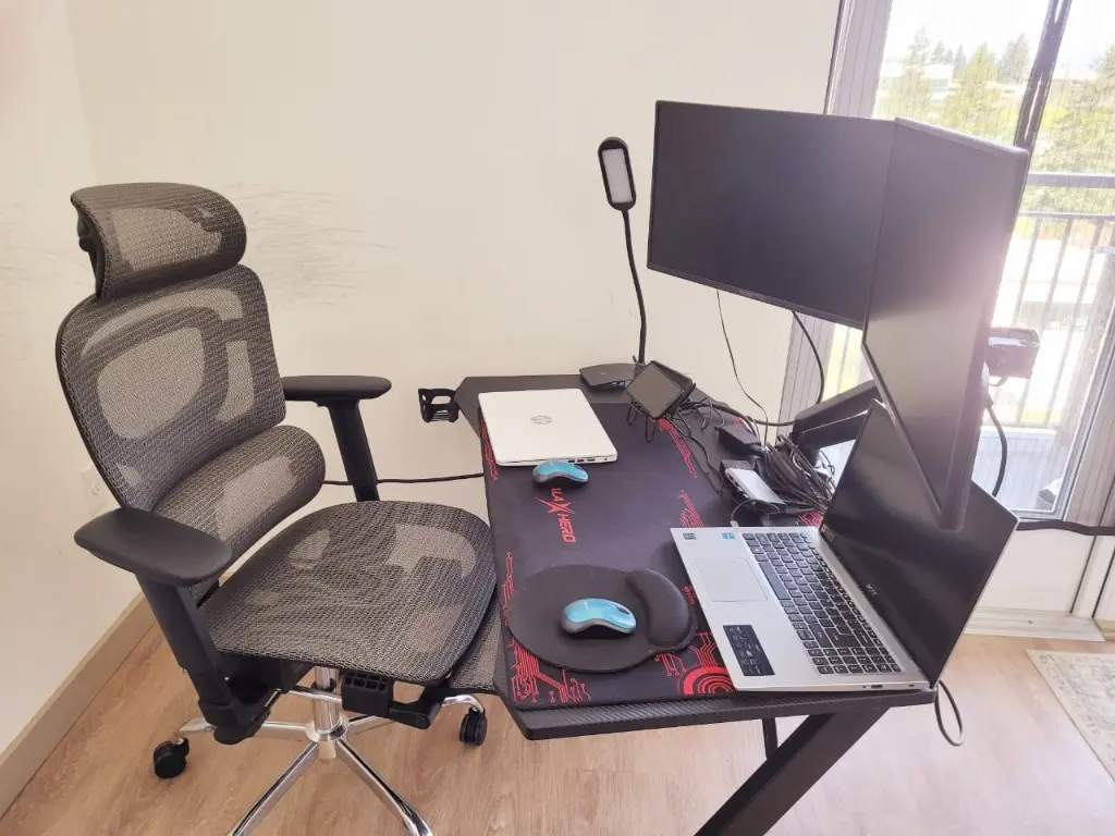 A mesh office chair with headrest and adjustable arms beside a desk with two monitors and a laptop.