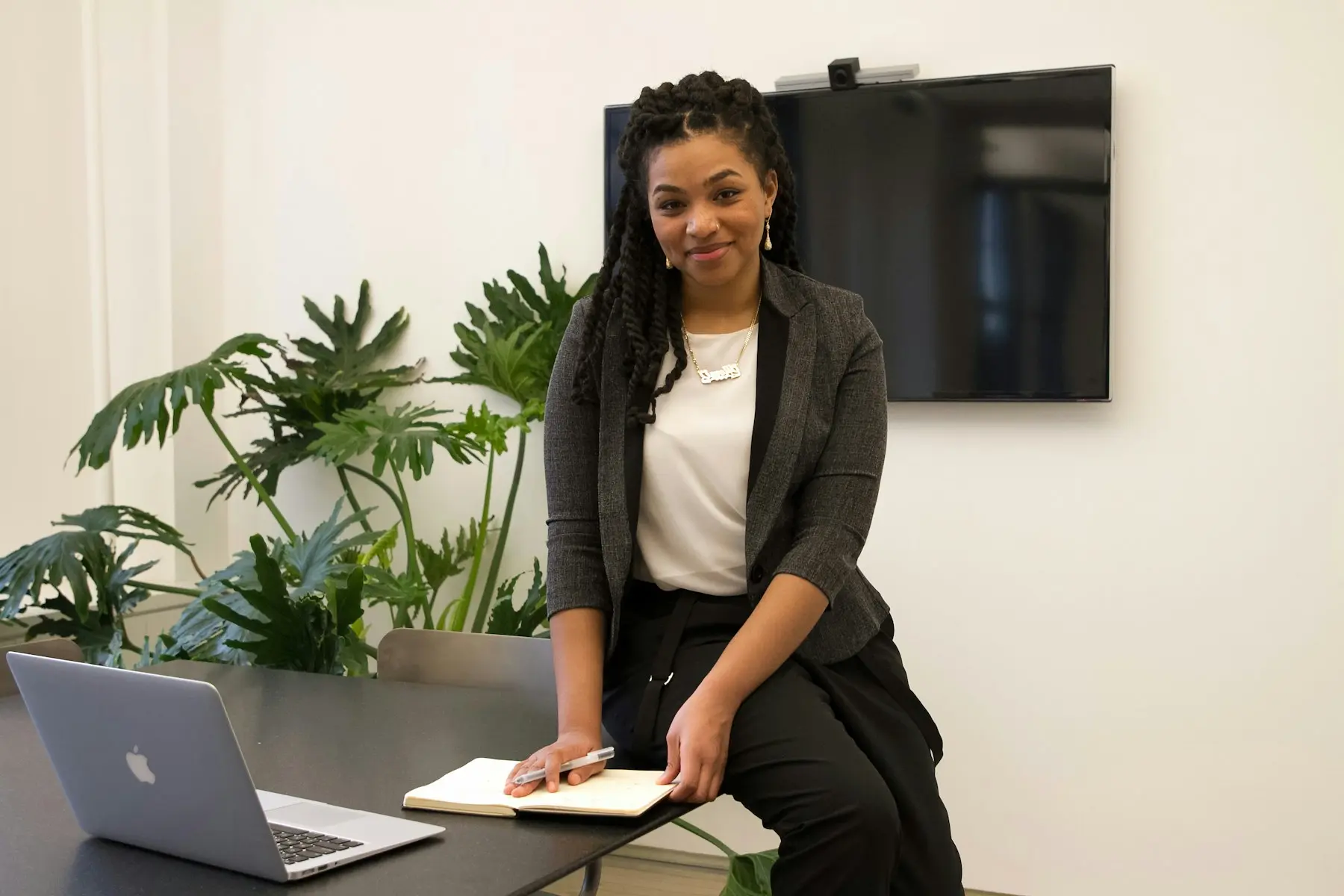 A professional standing at a desk with a laptop and notebook, beside a wall screen and plants.